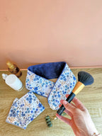 Bandeau de soin du visage en eponge de bambou bleu denim aux motif fleuris. Accompagner de lingettes et mini gant assortie une femme sapprete à realiser sa routine beauté et tient a la main un pinceau à maquillage.