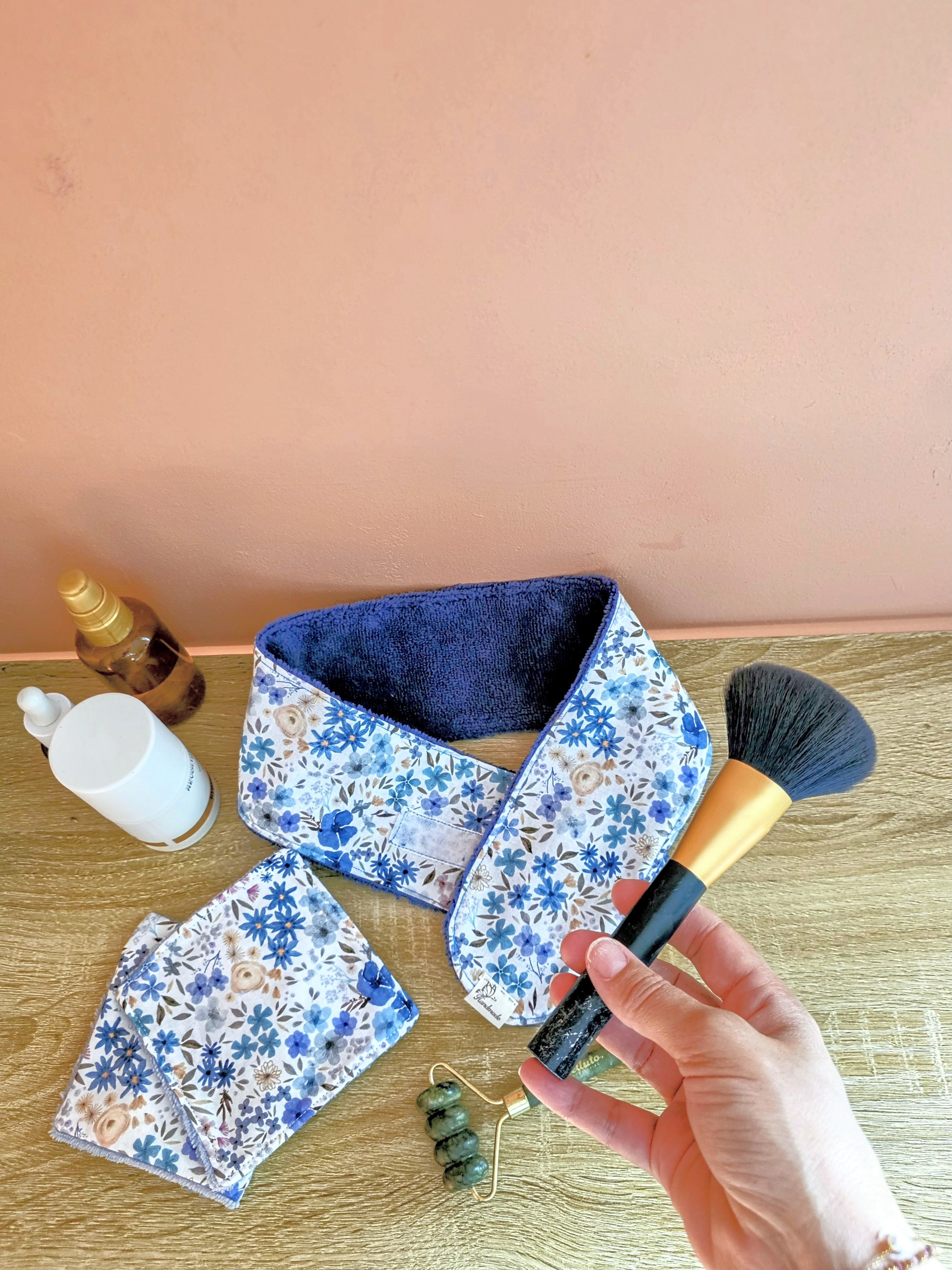 Bandeau de soin du visage en eponge de bambou bleu denim aux motif fleuris. Accompagner de lingettes et mini gant assortie une femme sapprete à realiser sa routine beauté et tient a la main un pinceau à maquillage.