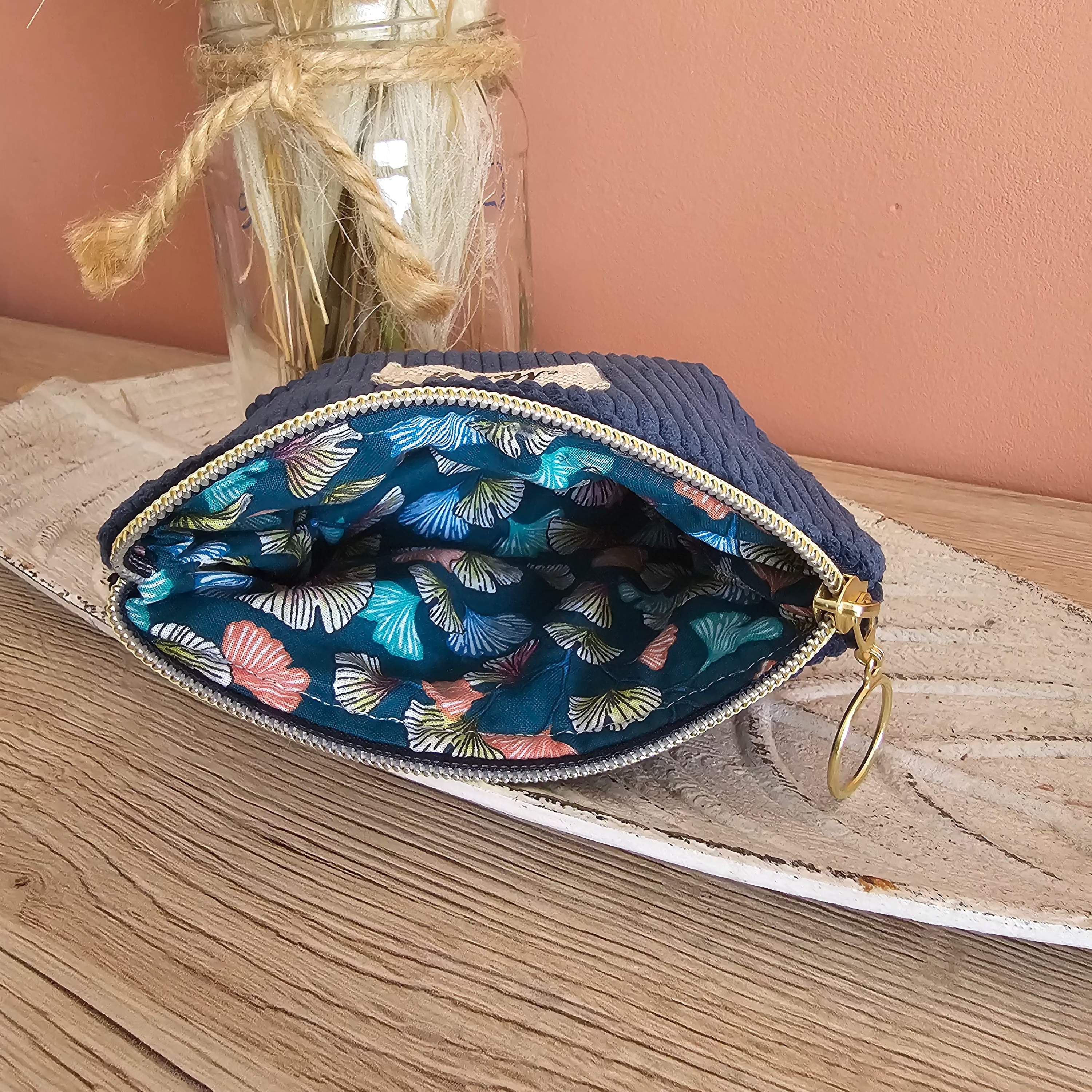 Vue sur l'intérieur de la mini pochette zippée qui est ouverte. Couleur bleu denim en velours à l'extérieur et coton bleu canard à motif ginkgo multicolores pastel à l’intérieur. Posé sur un meuble en bois avec une déco cocooning et douce de fleur sechée 