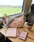 Sac bandoulière demi lune posé sur une table dans un train. Couleur velours bleuciel et coton aux motifs oiseaux de differente couleurs. Accompagné dun café a emporté et dun carnet de note et stylos.Ambiance cosy 