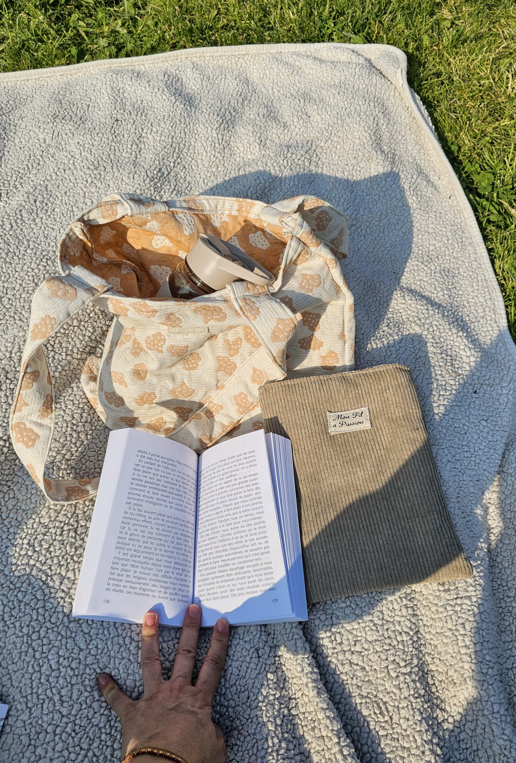 Une jeune femme posé sur un plaid dans un parc avec son tote bag, un livre et pochette à livre Mon fil à passion.  Une gourde fait partie du contenue du sac