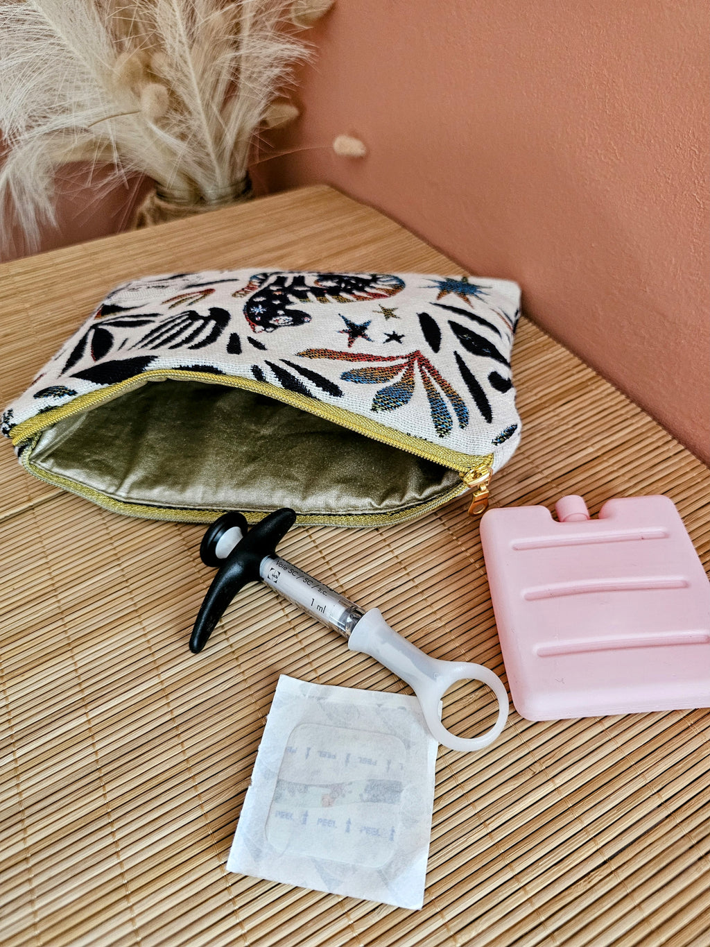 Pochette posée sur une table. Contient un médicament type piqûre injection qui nécessite d'être conserver au frais lors des transports ou voyage. Un bloc de glace et une compresse désinfectante. Trousse isotherme médicale au couleur beige et bleu et motif mystique. Coton enduit doré.