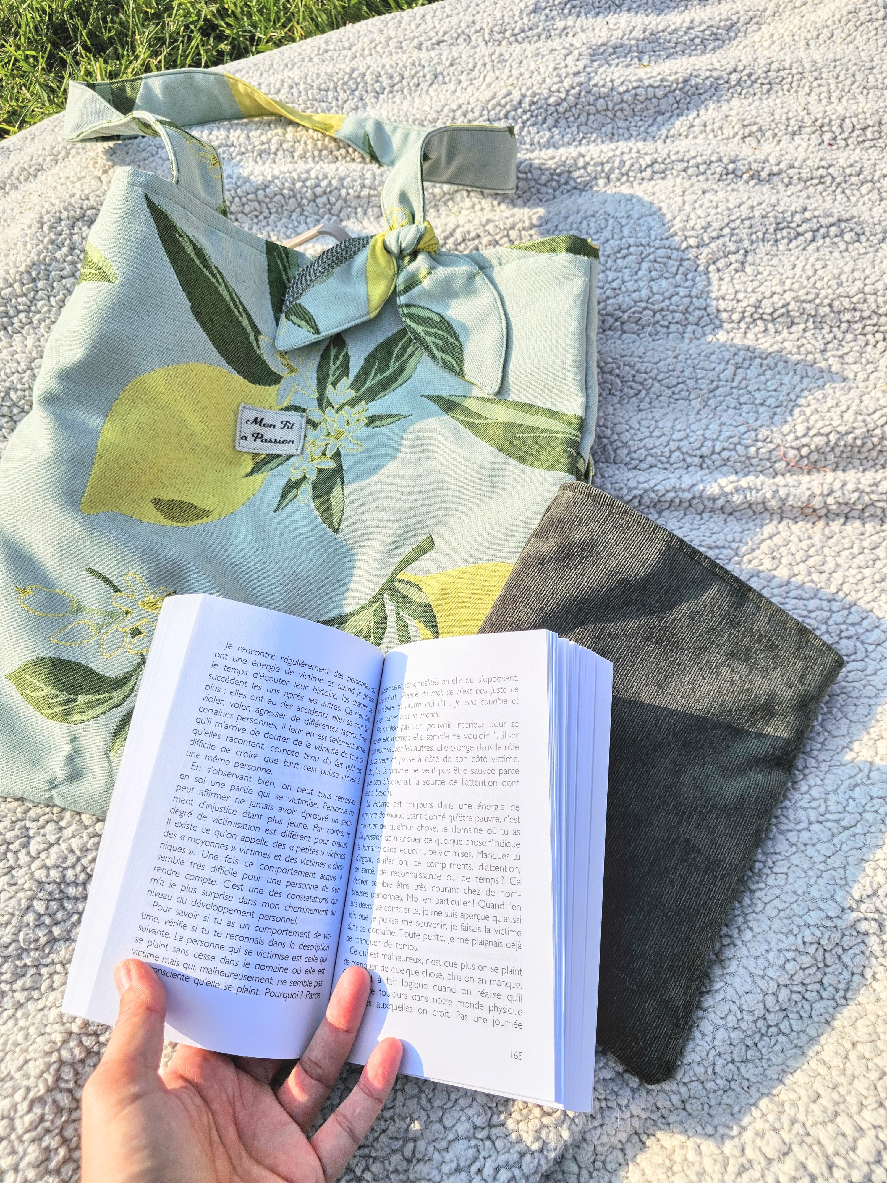 Femme installé sur une couverture de pique nique avec un livre et son tote bag citron contient une gourde et une housse protectrice pour livre Mon Fil à Passion 