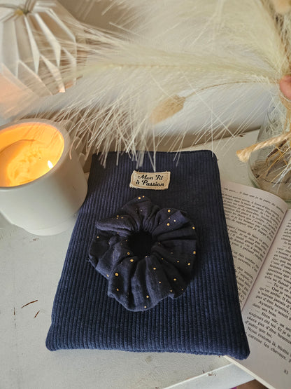 Pochette à livre artisanale en velours bleu marine. Pochette molletonnée. La pochette est posée sur un livre ouvert et un chouchou assorti en gaze de coton bleu marine et doré complète l’ensemble 